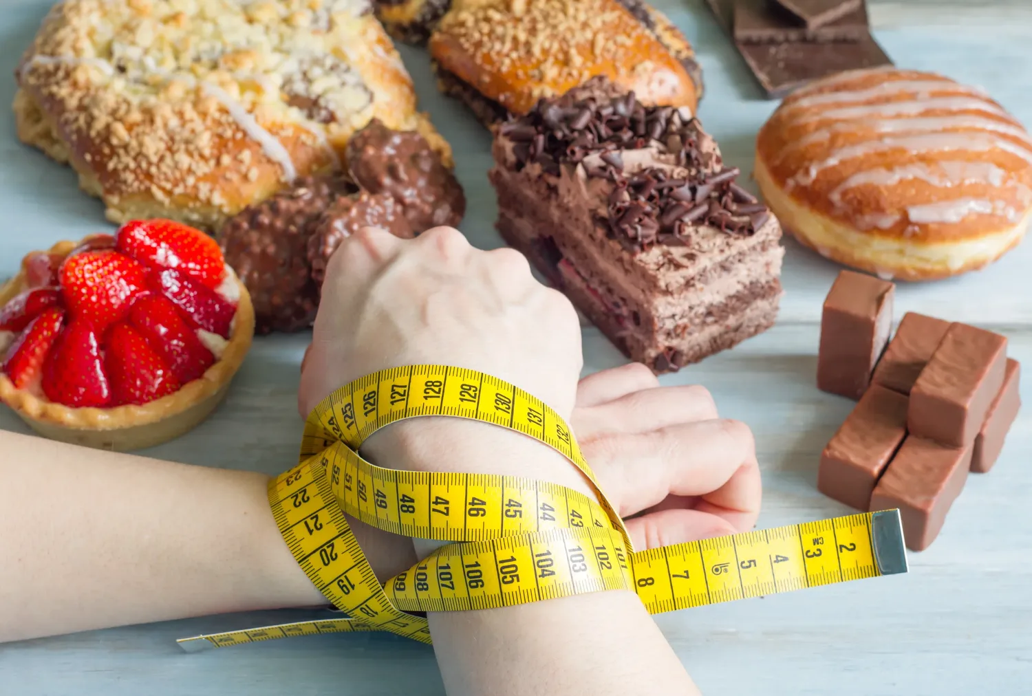 A measuring tape wrapped around a person's wrists in front of a table of pastries and cakes, illustrating the pressure of restrictive dieting.